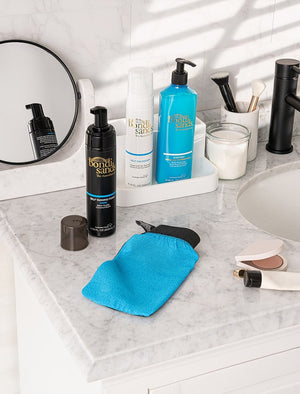 A marble bathroom countertop displays BondiSandsEU Self Tanning Foam Dark, a blue mitt, makeup items, a round mirror, candle, and black soap dispensers. Sunlight casts bronzed shadows across the neatly arranged scene.