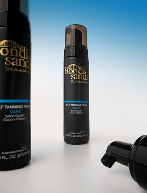 A bottle of BondiSandsEU Self Tanning Foam Dark stands upright on a white surface, with another bottle on the left and a pump nozzle on the right, set against a gradient blue-to-white background for a flawless bronzed glow.