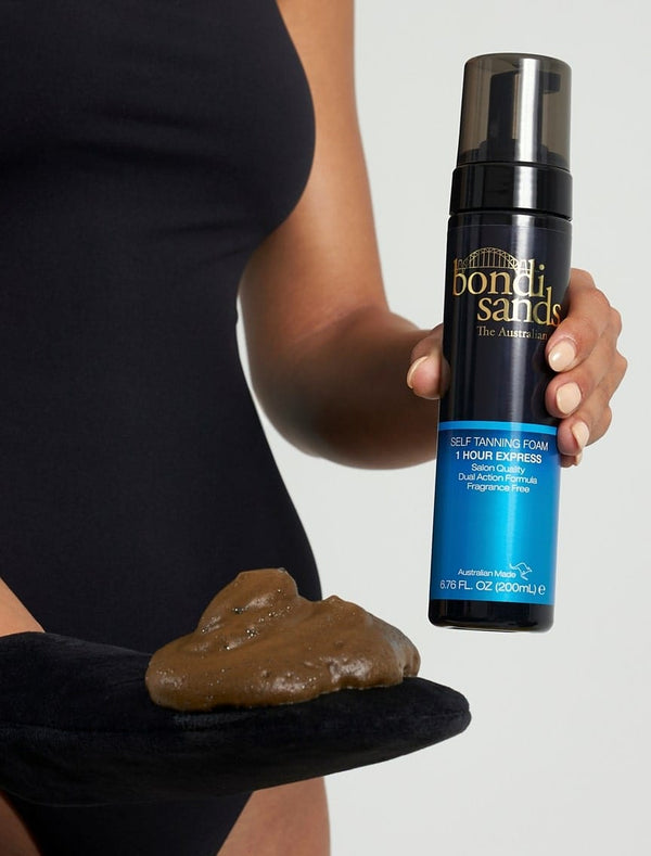 A person in a black sleeveless outfit holds a bottle of BondiSandsEU Self Tanning Foam 1 Hour Express in one hand and a tanning mitt with foam in the other.