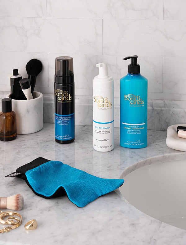 Three BondiSandsEU self tanning foams, including the Self Tanning Foam 1 Hour Express, are shown with a blue mitt on a marble bathroom counter near a sink, surrounded by makeup brushes, bottles, and toiletries.