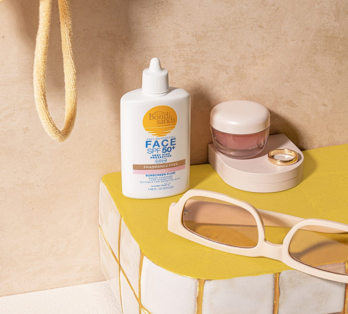 A bottle of BondiSandsEU Face SPF 50+ Tinted Everyday Sunscreen Fluid sits on yellow tiles beside a small jar, gold ring, and beige sunglasses, with a towel hanging in the background.