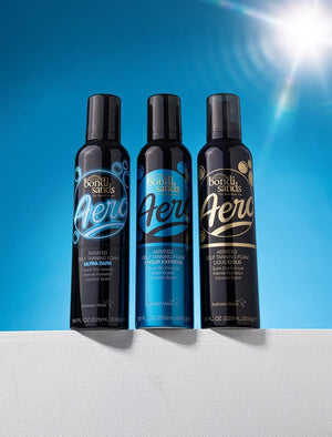 Three BondiSandsEU Aero Self Tanning Foam Liquid Gold bottles stand side by side against a bright blue sky, sunlight and lens flare illuminating their black design with blue and gold accents, promising a long-lasting, natural-looking tan.