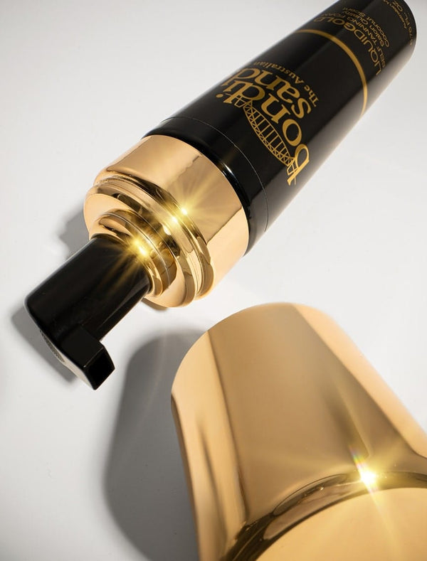 A black and gold bottle of BondiSandsEU Self Tanning Foam Liquid Gold with a pump dispenser rests on a white surface, its shiny accents and lighting evoking a luxurious liquid gold effect.