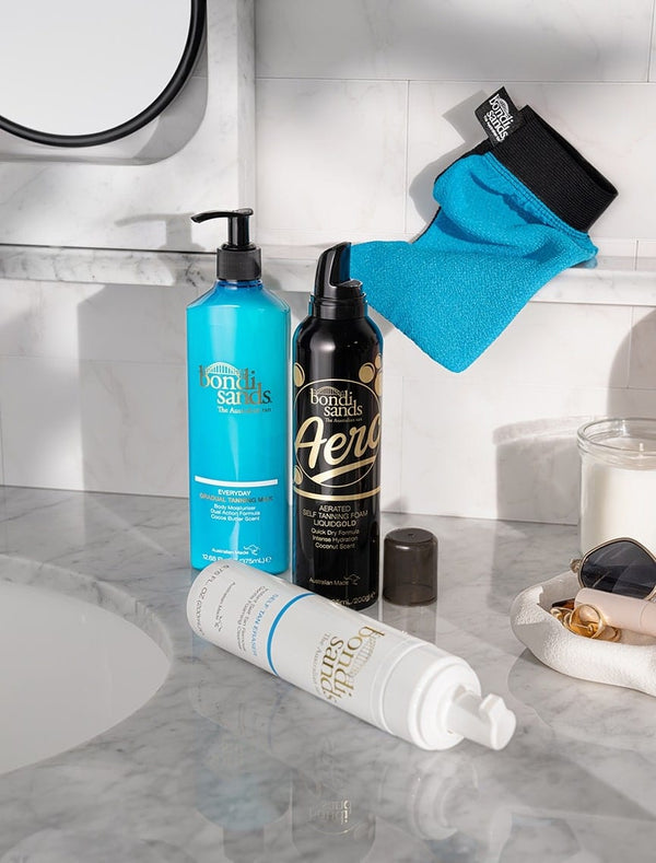 Three BondiSandsEU tanning products, including Aero Self Tanning Foam Liquid Gold, and a blue mitt rest on a marble counter with sunglasses, a candle, and hair clip. A mirror on the left hints at a long-lasting natural tan.