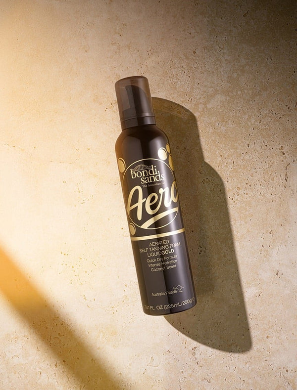 A black bottle of BondiSandsEU Aero Self Tanning Foam Liquid Gold sits on a sunlit beige surface, its gold and white label highlighting Ultra Dark and Coconut Scent for a long-lasting, natural-looking tan.
