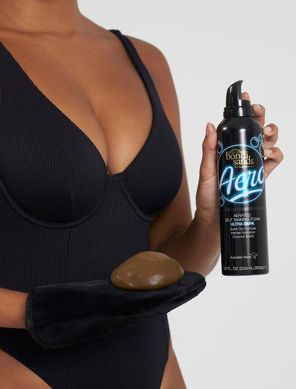 Wearing a black swimsuit, a person holds BondiSandsEU Aero Self Tanning Foam Ultra Dark in one hand and a tanning mitt with foam in the other, poised to achieve a flawless bronzed tan against a plain backdrop.