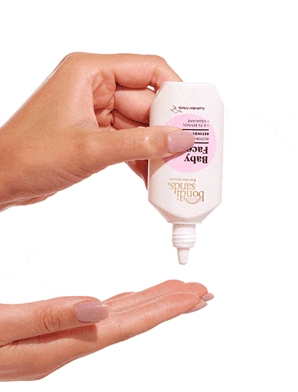 A hand squeezes a white bottle of BondiSandsEU Baby Face Retinol Serum, dispensing serum onto the palm of another hand against a white background, helping protect and maintain youthful-looking skin.
