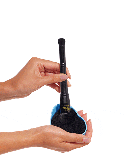 A hand cleans a makeup brush on a black sponge in a blue container, showing how to prep brushes for flawless application of the BondiSandsEU Self Tan Contour Kit against a white background.