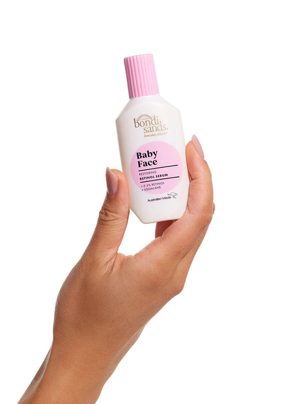 A hand holds a BondiSandsEU Baby Face Retinol Serum bottle with a pink cap and label, set against a white background—ideal for targeting fine lines and promoting youthful-looking skin.