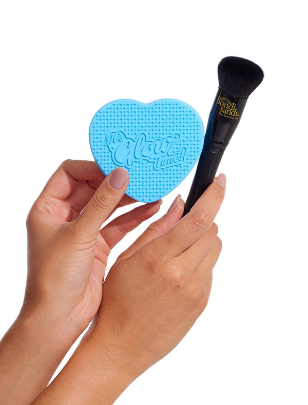 Two hands hold the BondiSandsEU Self Tan Contour Kit, featuring a blue heart-shaped cleaning pad and a black makeup brush—ideal for flawless bronzing or contouring—set against a clean white background.