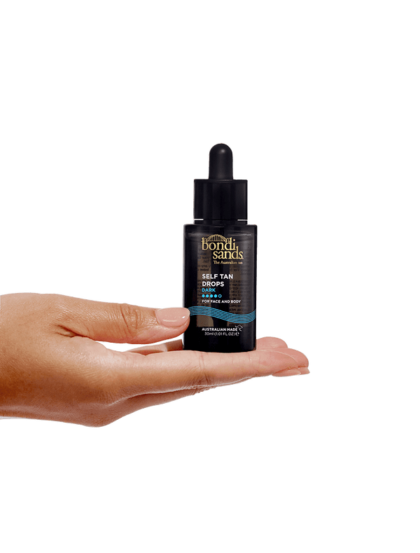 A hand holds a black bottle of BondiSandsEU Self Tan Drops Dark with a dropper lid against a plain white background, showcasing these Australian self tanning drops.