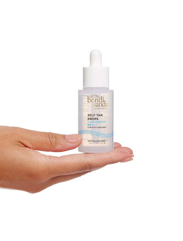 A hand holds a clear bottle of BondiSandsEU Self Tan Drops Light/Medium with a white dropper cap. The label has gold and blue text, promising glowing skin and an Australian tan for face and body against a white background.