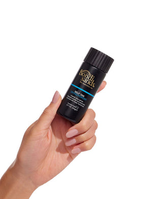 A hand with manicured nails holds a small black bottle of BondiSandsEU Self Tan Drying Powder against a white background, highlighting an essential step in achieving a flawless tan.