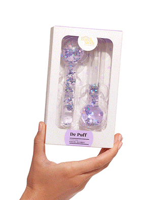 A hand holds a BondiSandsEU De Puff Cooling Facial Globes box, showing purple glitter-filled globes through a window. The mainly white packaging has purple and gold accents and helps reduce puffiness.