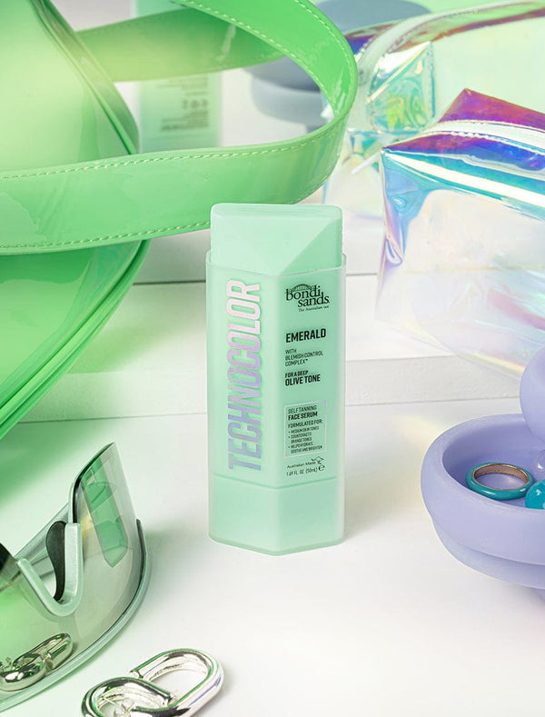 A green bottle of BondiSandsEU Technocolor Emerald Self Tanning Face Serum for medium skin tones stands upright amid green sunglasses, a translucent green bag, iridescent pouches, a blue ring dish, and a metallic clasp on a white surface.