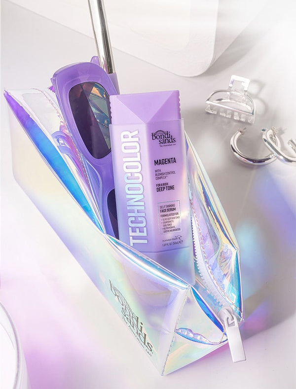 A holographic makeup bag holds purple sunglasses and a tube of BondiSandsEU Technocolor Magenta Self Tanning Face Serum, with reflective accessories placed nearby on a white surface.