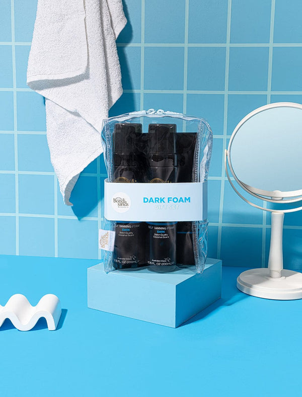 A clear toiletry bag with black BondiSandsEU Dark Foam Gift Set bottles sits on a blue pedestal in a blue-tiled bathroom, with a white towel, round mirror, white décor piece, and Application Mitt nearby.