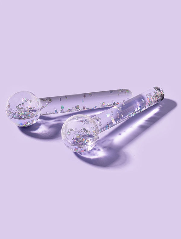 The BondiSandsEU De Puff Cooling Facial Globes—clear, glitter-filled wands with round ends—rest on a pastel purple surface. They help reduce puffiness and support lymphatic drainage while adding a touch of shimmer.