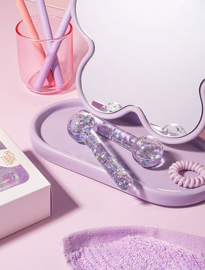 Two BondiSandsEU De Puff Cooling Facial Globes with glitter rest on a lavender tray beside a mirror. A pink coil hair tie, purple towel, and straws complete the setup—great for lymphatic drainage or reducing puffiness.