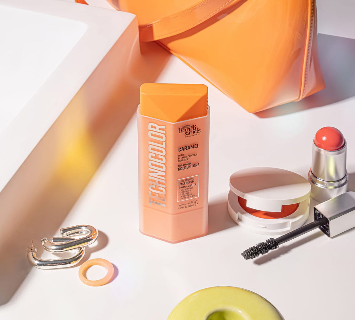 A bottle of BondiSandsEU Caramel Technocolor Self Tanning Face Serum sits on a white surface beside a coral handbag, earrings, an orange ring, coral blush for deep skin tones, a mascara wand, and a green donut-shaped item for a natural glow.