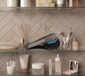 A bathroom shelf holds toiletries, makeup brushes, a comb, and the BondiSandsEU Back Applicator for a smooth tan. A reed diffuser adds style, while light herringbone tiles provide an elegant backdrop.