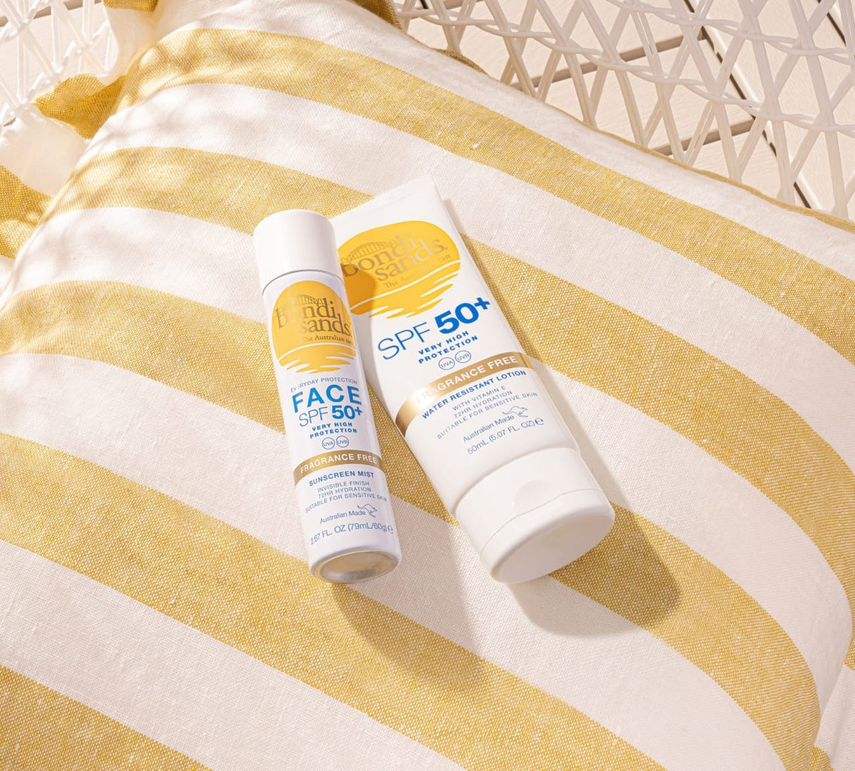 A BondiSandsEU SPF 50+ Everyday Sunscreen Face Mist and lotion sit on a white-yellow striped cushion, capturing a summery vibe. Enjoy fast-absorbing protection against UVA & UVB as sunlight casts soft shadows.