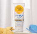 A tube of BondiSandsEU SPF 50+ Everyday Body Sunscreen Lotion stands upright on a white surface with a yellow towel and blue sunglasses nearby, sheer white curtains in the background—ideal for daily body sun protection.