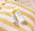 Two bottles of BondiSandsEU SPF 50+ Everyday Sunscreen Face Mist, providing UVA & UVB protection, sit on a white and yellow striped pillow with a woven chair behind, all beautifully illuminated by natural sunlight.