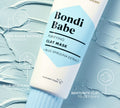 A tube of BondiSandsEU Bondi Babe Clay Mask rests on blue clay, featuring red algae, blue spirulina, and bentonite clay to detoxify skin and reveal a glowing, healthy radiance.