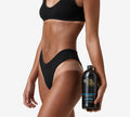 A person in a black bikini displays a distinct tan line on their hip while holding BondiSandsEU’s Dark Salon Solution 1L Duo against a plain background.