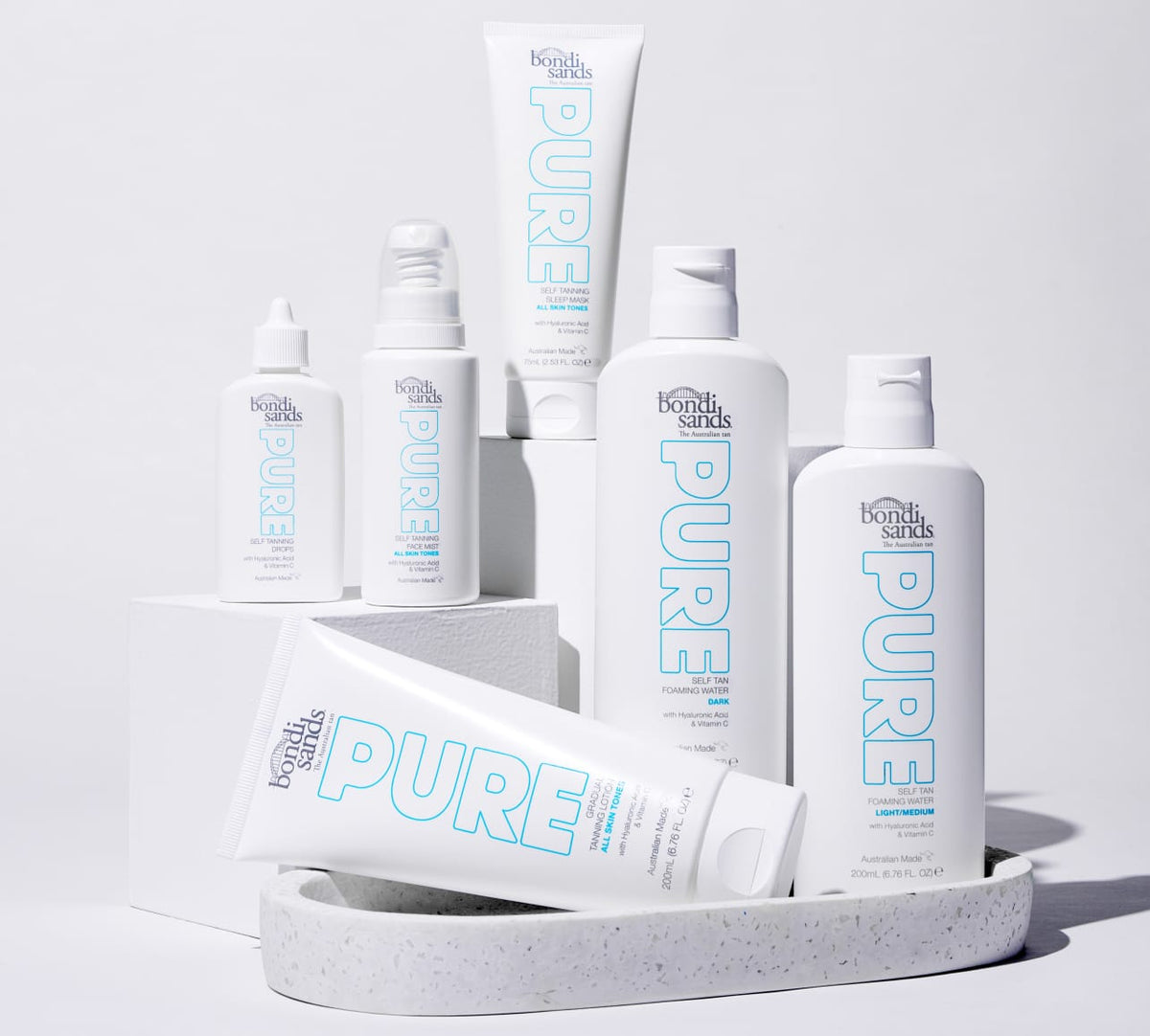 Several white bottles and tubes of BondiSandsEU’s Pure Self Tanning Sleep Mask are neatly displayed on a white tray and blocks with light blue lettering, set against a clean white background.