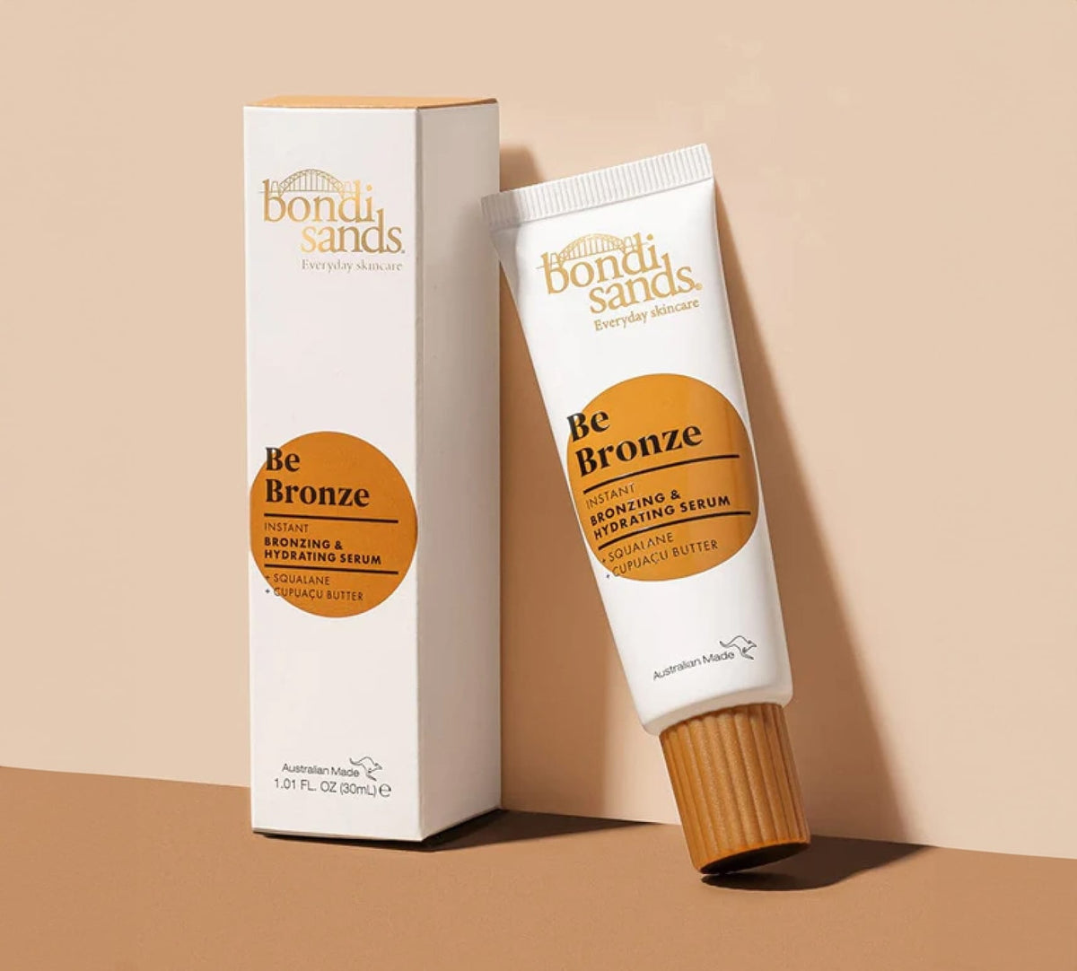 A tube and box of BondiSandsEU Be Bronze Instant Bronzing & Hydrating Serum are displayed on a beige background. The white packaging has orange accents, text, and a screw-on cap, highlighting this hydrating, glow-boosting serum.