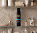 A BondiSandsEU Dark Foam Gift Set rests on a glass shelf, surrounded by makeup brushes, skincare bottles, and beauty products against a tiled bathroom wall.