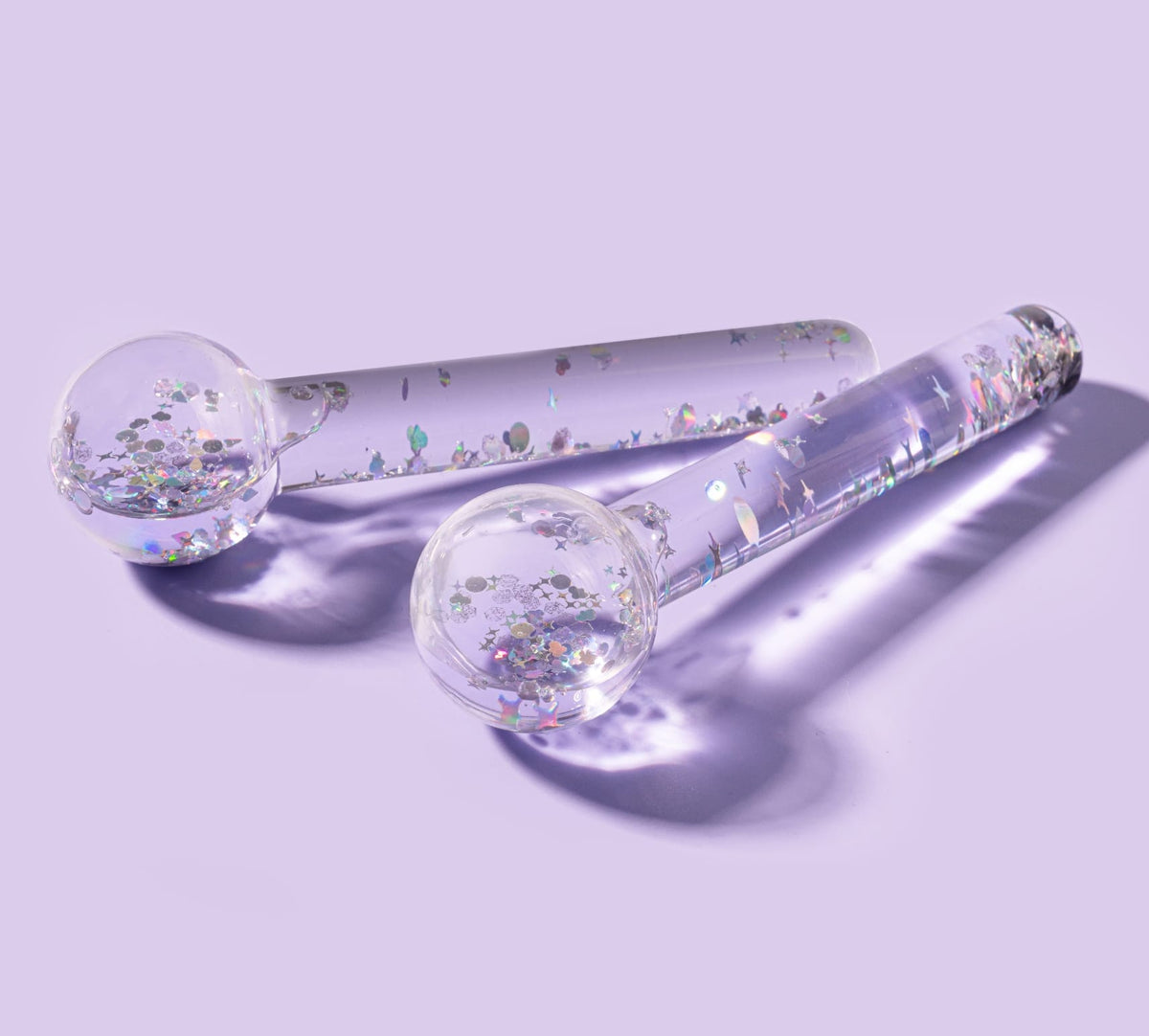 The BondiSandsEU De Puff Cooling Facial Globes—two clear glass wands with colorful glitter and shapes—rest on a pale lavender surface, casting soft shadows, ideal for de-puffing and lymphatic drainage routines.