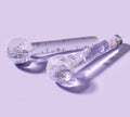 The BondiSandsEU De Puff Cooling Facial Globes—two clear glass wands with colorful glitter and shapes—rest on a pale lavender surface, casting soft shadows, ideal for de-puffing and lymphatic drainage routines.