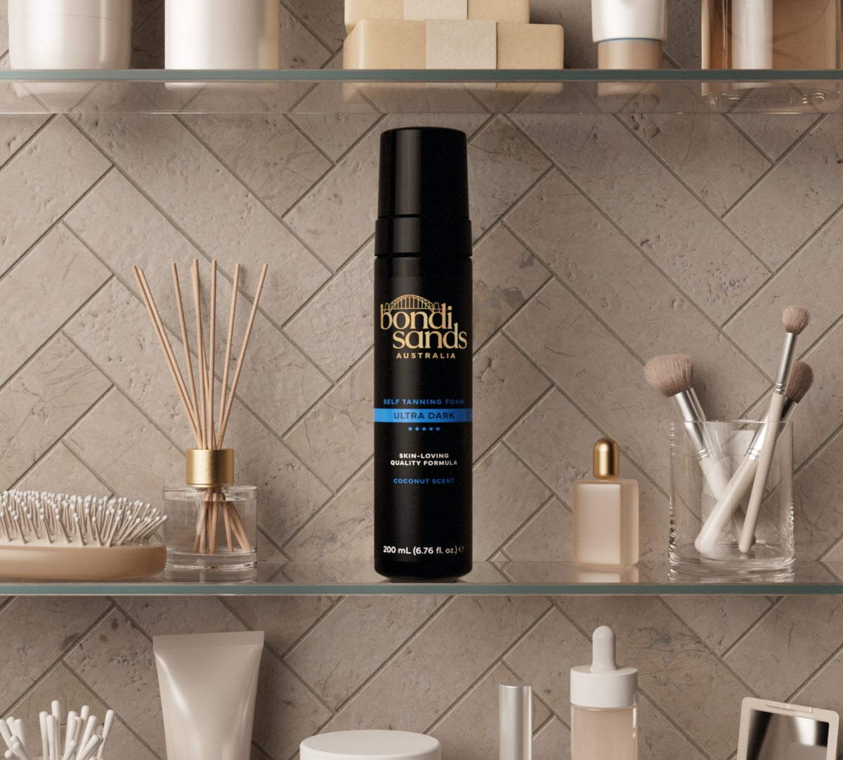 A bottle of BondiSandsEU Ultra Dark Self Tanning Foam rests on a bathroom shelf among makeup brushes, perfume bottles, and decorative accessories, set against a tiled wall.
