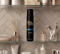 A bottle of BondiSandsEU Ultra Dark Self Tanning Foam rests on a bathroom shelf among makeup brushes, perfume bottles, and decorative accessories, set against a tiled wall.