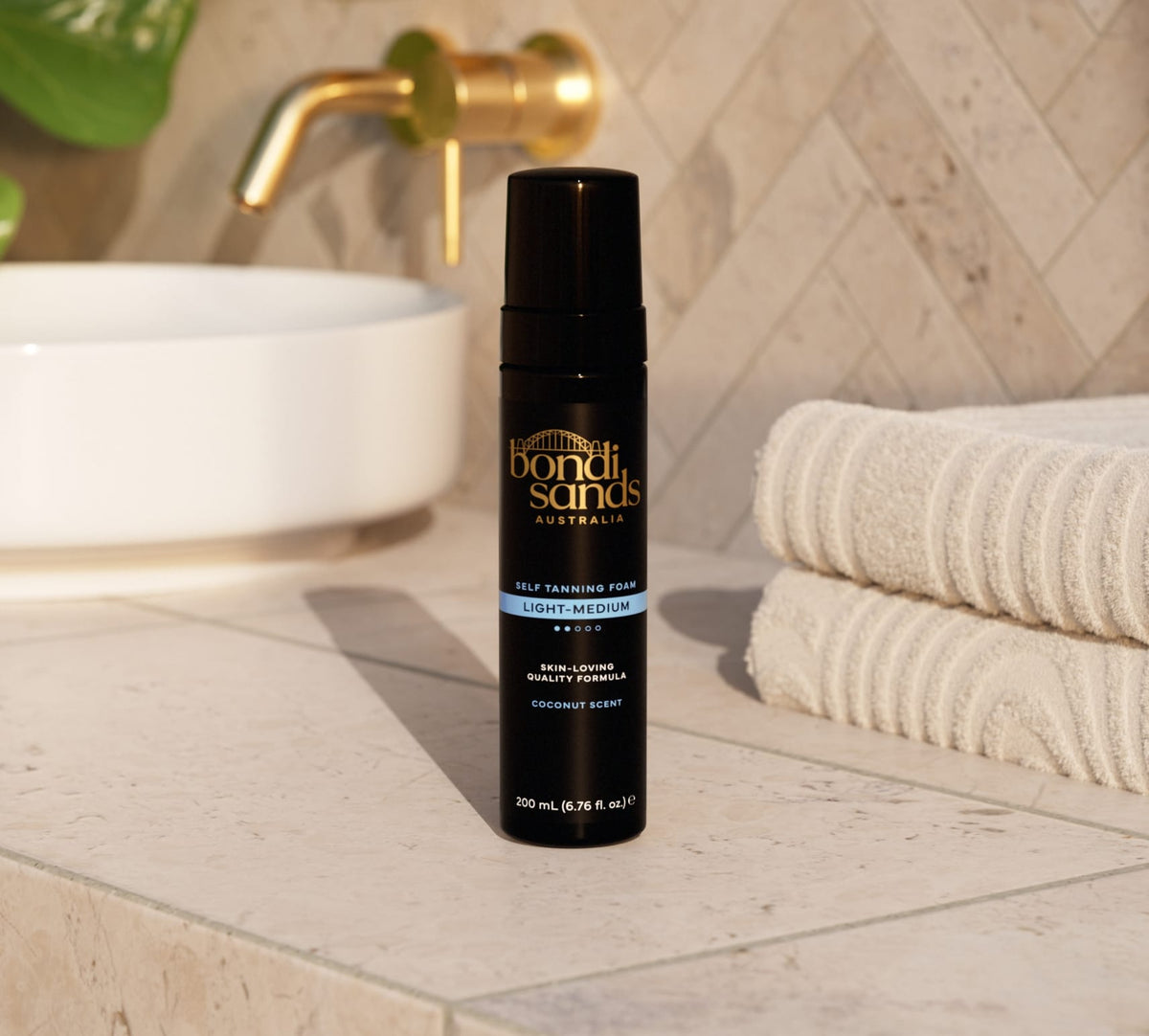 A BondiSandsEU Light/Medium Self Tanning Foam & Application Mitt Bundle rests on a bathroom counter next to folded white towels, sunlight highlighting the products to suggest a flawless, natural glow.