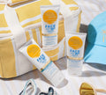 Three tubes of BondiSandsEU SPF 50+ Everyday Matte Tinted Face Lotion sit on a yellow and white striped bag with a blue cap, sunglasses, and sand—perfect for summer days and great for shine-free sun protection.