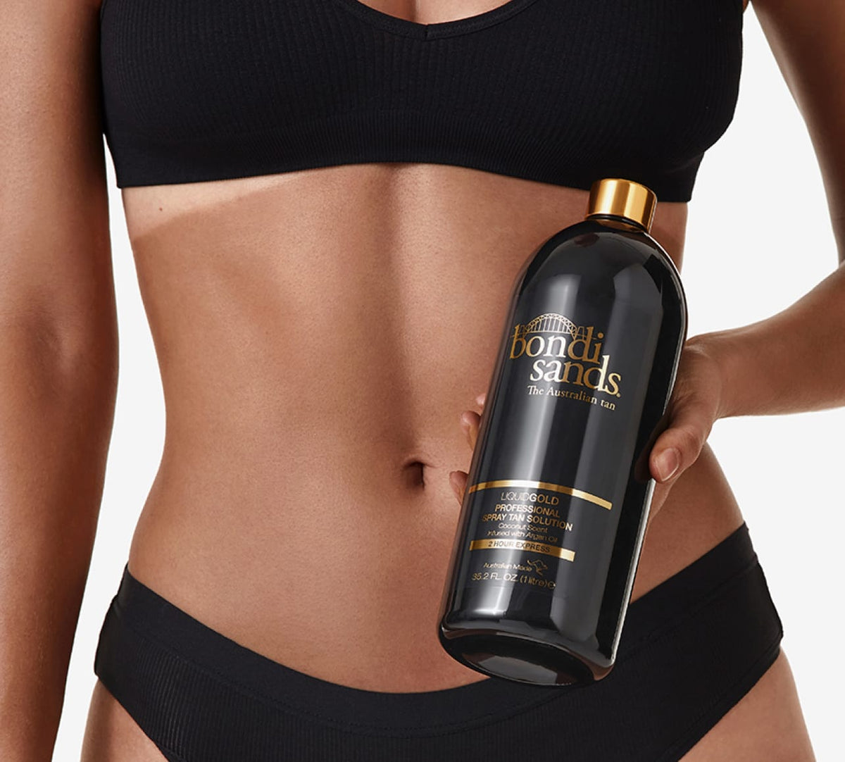 A person in black underwear holds a large black and gold BondiSandsEU Liquid Gold Salon Solution Bundle bottle against their bare stomach.