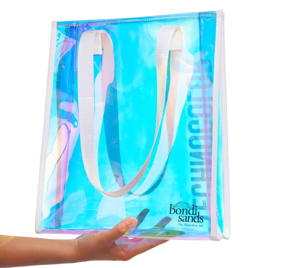 A hand holds the Technocolor Tote Bag by BondiSandsEU, a transparent iridescent bag with white handles and a Bondi Sands logo in the lower right corner. The background is white.