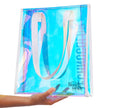 A hand holds the Technocolor Tote Bag by BondiSandsEU, a transparent iridescent bag with white handles and a Bondi Sands logo in the lower right corner. The background is white.
