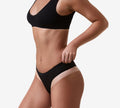 A woman in matching black bralette and black underwear with beige trim, seen from the side on a light background, displays glowing skin as if enhanced by BondiSandsEU Salon Solution Light-Medium, a fast-drying professional spray tan.