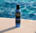 A blue can of BondiSandsEU Express Aero Self Tanning Mousse stands upright on a stone surface by a pool, promising a quick-dry, deep bronze glow against the sparkling blue water in the background.