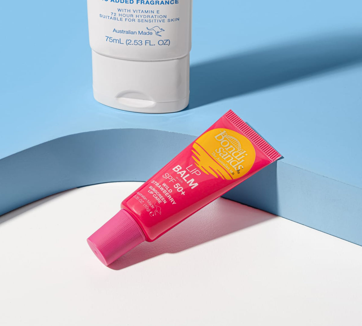 A pink tube of BondiSandsEU Strawberry SPF 50+ Lip Balm sits on a white surface, with a white bottle partly visible in the background against a blue and white backdrop.