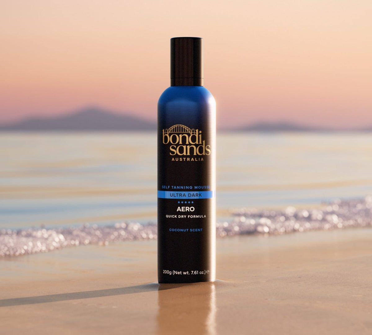 A BondiSandsEU Ultra Dark Aero Self Tanning Mousse stands upright on wet sand by the shore, with gentle waves and distant mountains in golden light—showcasing its quick-dry formula for a flawless glow.