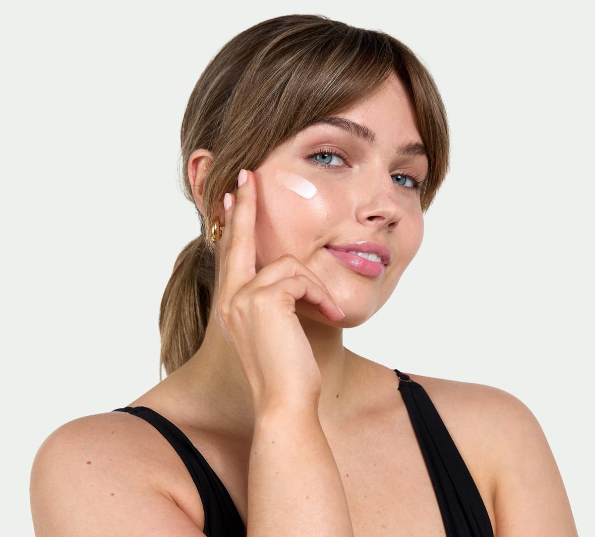 A woman with light brown hair in a ponytail applies BondiSandsEU Gradual Self Tanning Face Lotion to her cheek, smiling gently. She wears a black sleeveless top and gold hoop earrings against a plain, light background.