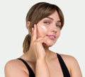 A woman with light brown hair in a ponytail applies BondiSandsEU Gradual Self Tanning Face Lotion to her cheek, smiling gently. She wears a black sleeveless top and gold hoop earrings against a plain, light background.