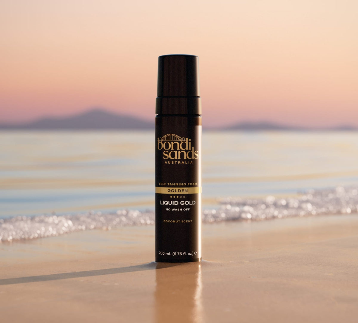A BondiSandsEU Glow & Go Bundle sits upright on sandy beach with waves and distant mountains, offering a golden tan and transfer-resistant finish in warm sunlight.