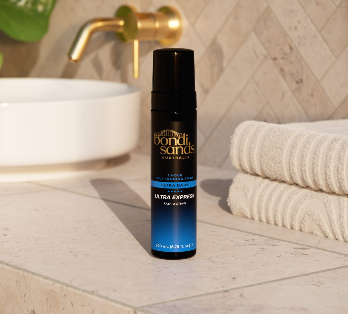 A bottle of Bondi Sands Europe Ultra Express Self Tanning Foam sits on a bathroom counter beside folded white towels, with a white sink and gold faucet in the background.